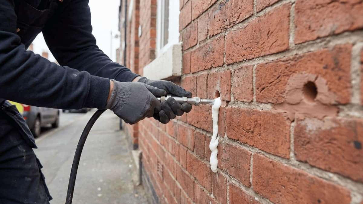Een vakman in donkere werkkleding en grijze handschoenen drukt een metalen injectiepistool tegen een boorgat in een rode bakstenen gevel terwijl wit isolatieschuim naar buiten druipt.
